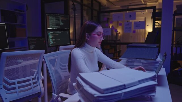 Tired Asian woman sitting at her desk surrounded by stacks of paperwork, reviewing details and working overtime in the office at night.