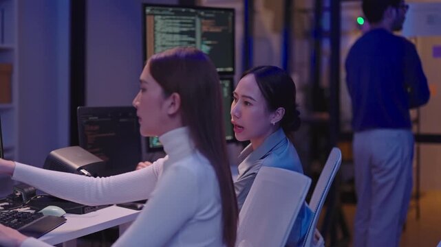 Asian women colleagues collaborating and talking about software development projects in a night office environment.