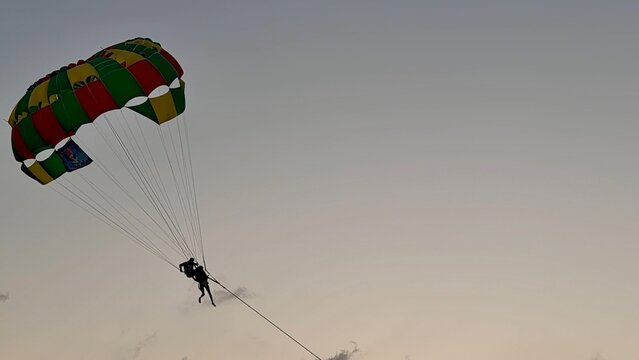 Silhouette of a person parasailing high in the sky over the ocean during a golden tropical sunset. Scenic summer vacation activity with bright sun, orange clouds, and serene sea horizon.