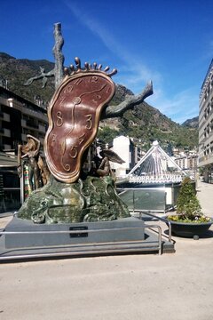 The Nobility of Time bronze sculpture by Salvador Dali. A melting clock on a tree trunk monument with mountains and the Paris Bridge in the background. Andorra la Vella, Andorra - May 7, 2017