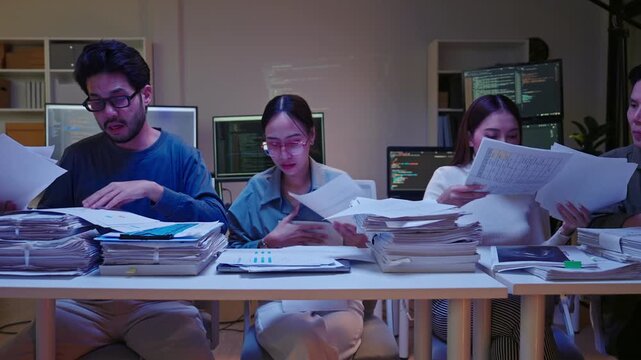 Group of Asian colleagues pan shot searching through disorganized piles of documents, feeling rushed and stressed to meet a night deadline.