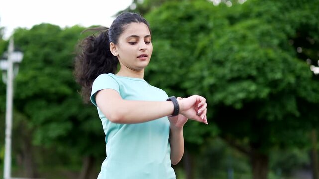 Young female runner jogs through an urban park while checking her smartwatch. Fit sportswoman monitors her fitness progress and enjoys running outdoors, staying active and healthy in city environment.