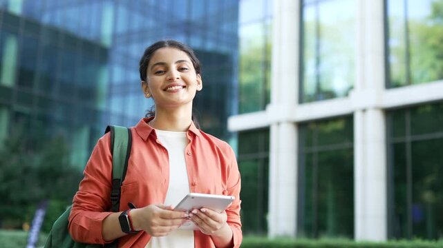 Portrait of a happy female student using a digital tablet while standing on campus near a university building, then looking at the camera with a confident smile. Young woman poses outdoors, close up.