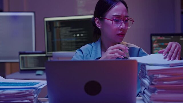 Close up of a frustrated Asian female employee rubbing her eyes and forehead, feeling burned out while searching through documents late at night.