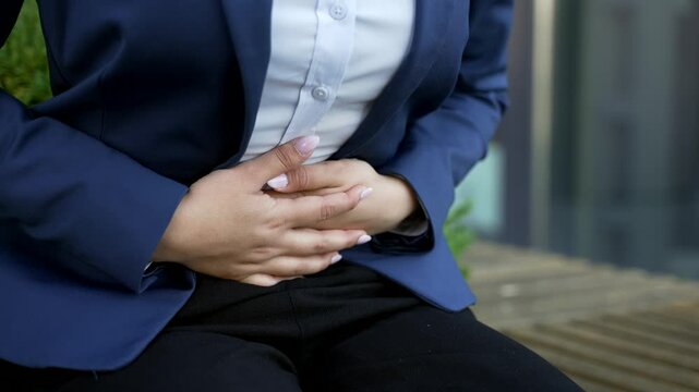 Close up of female hands holding and massaging the stomach as a businesswoman in suit feels abdominal pain sitting on bench outside. Female employee experiences cramps, discomfort, spasms, constipated