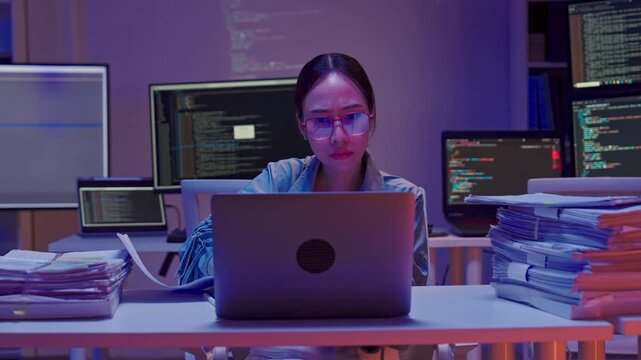 Tired Asian woman sitting at her desk surrounded by stacks of paperwork, reviewing details and working overtime in the office at night.