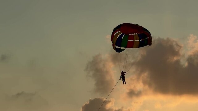 Parasailing activity over the sea at sunset on Phuket island, Thailand. Tourist flying with colorful parachute above the ocean with boats and jet skis on the shore during golden hour.