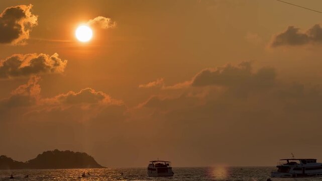 Parasailing activity over the sea at sunset on Phuket island, Thailand. Tourist flying with colorful parachute above the ocean with boats and jet skis on the shore during golden hour.