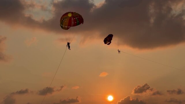 Parasailing activity over the sea at sunset on Phuket island, Thailand. Tourist flying with colorful parachute above the ocean with boats and jet skis on the shore during golden hour.