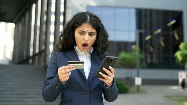 Upset businesswoman notices unauthorized charges while checking her credit card on smartphone near an office building. Female employee reacts to a financial problem with anxiety and concern, close up.