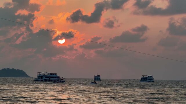 Parasailing activity over the sea at sunset on Phuket island, Thailand. Tourist flying with colorful parachute above the ocean with boats and jet skis on the shore during golden hour.