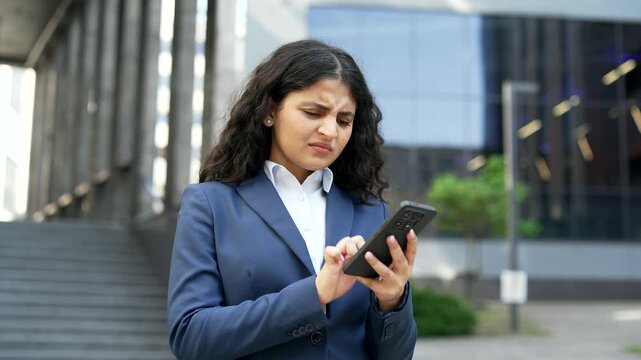 Confused businesswoman struggles while using her smartphone near an office building. Female employee experiences difficulty with mobile task, frustration and uncertainty in urban environment, close up