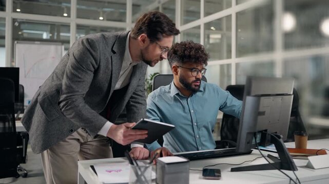 Two diversity business men talking reviewing working tasks in digital tablet and computer desktop monitor solving corporate problem talk Caucasian man boss help with work to Arabian intern in office