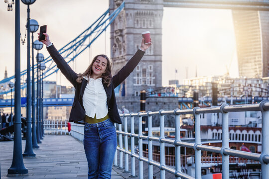 A happy, young woman walks on the street holding a mobile phone and a coffee cup in front of the urban city skyline of London
