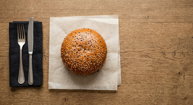 Overhead top view of a classic sesame seed burger bun on white parchment paper with cutlery and a black napkin on a wooden table, minimalist food composition with copy space for design
