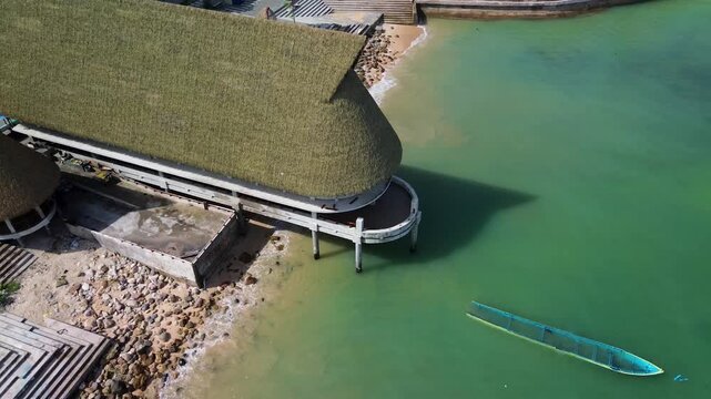 Aerial orbit of thatched roof overwater pavilion with wooden pier and traditional boat in turquoise tropical sea
