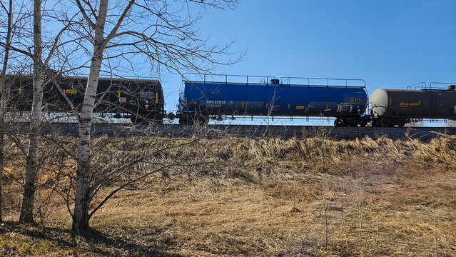 Freight train in early Spring with UTLX and SHPX tank cars passing through a rural landscape under a blue sky (Anoka County, MN - March 29, 2026). For Editorial Use Only.
