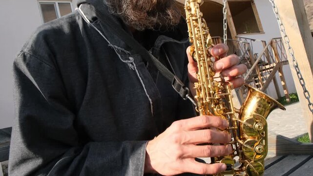Mysterious and romantic street musician playing golden saxophone sparkling in sunlight. Bearded street musician sitting on swing near mansion performing melody. Bearded musician in hat alone