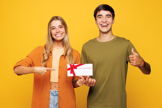 Young couple two friends family man woman wear casual clothes look camera together hold point on gift certificate coupon voucher card for store isolated on pastel plain light yellow color background.
