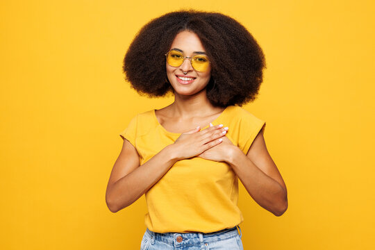 Young grateful smiling happy woman of African American ethnicity she wear t-shirt casual clothes put folded hands on heart isolated on plain yellow orange background studio portrait Lifestyle concept