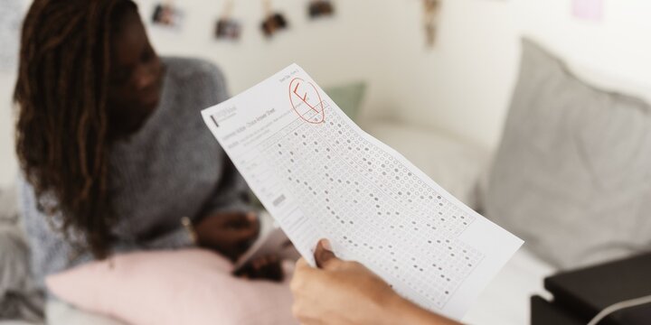 A person holds a test paper with a failing grade. In the background, another person sits on a bed. The focus is on the test paper and the failing grade. Education concept.