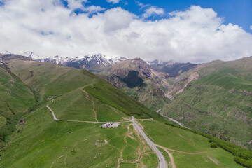 Naklejka premium Aerial View of Kazbegi Mountains in Georgia, Winding Road and Green Hills Landscape, Snow-Capped Caucasus Peaks. Adventure Travel and Tourism Concept, Majestic Nature Scene with Clouds and Sky