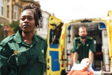 Emergency responders assist a patient. A Black female paramedic stands confidently. A male paramedic pushes a stretcher. Ambulance in the background. Healthcare concept. © Rawpixel.com