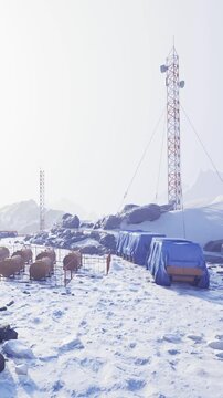 A group of storage containers and equipment is set up at a research station in a snowy area. Mountains are visible in the background under a clear sky. The scene shows a cold, remote environment.