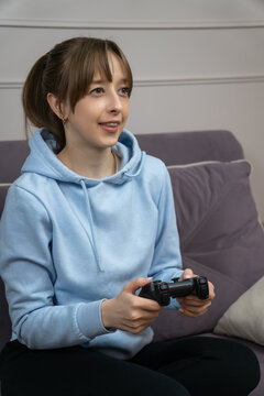 Gamer girl sitting on comfortable sofa holding wireless controller, enjoying playing video game, looking at screen and smiling, having fun at home. Vertical photo