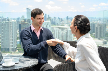 A businessman and a businesswoman dressed casually shake hands after closing a deal on a rooftop overlooking the city skyline.