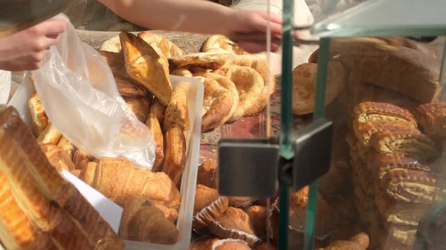 Baker uses metal tongs to pick and place fresh sweet traditional pastries into bag for customer. Pastry bakery filled with sunlight. Selling baked goods and bread. Bakery business