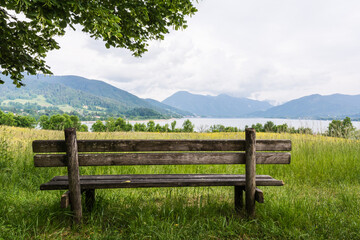 Fototapeta premium Empty wooden bench on a green meadow with a scenic view of Tegernsee lake and Alps