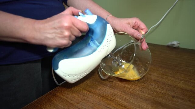 Hands mix eggs in a transparent bowl using a hand mixer on a wooden table.
