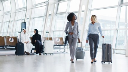 Caucasian attractive businesswoman in business style and her multiracial colleague walking through the corridor of the airport with suitcases, speaking and waiting for the plain