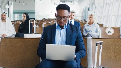 Multiracial businessman in a suit looking at the laptop and typing something while sitting at the departure hall and waiting for the plain. Traveling concept