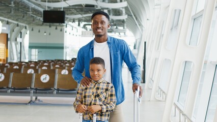 Portrait shot of the cute happy multiracial family of father and son standing at the airport with suitcase and smiling to the camera while waiting for their departure