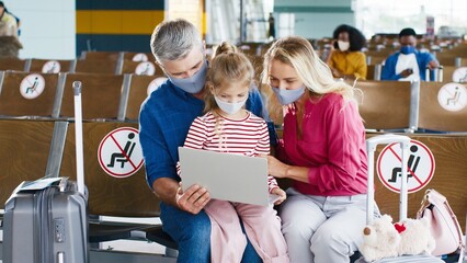 Cheerful family of parents and their daughter wearing protective masks watching something on the laptop computer while waiting for their departure in the airport