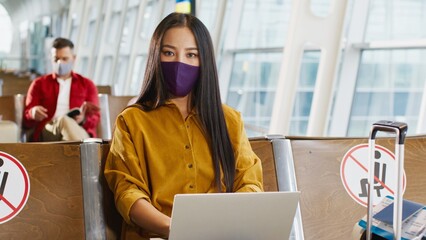 Asian young attractive woman with brunette hair wearing protective mask watching something or working on the laptop computer while waiting for her departure in the airport