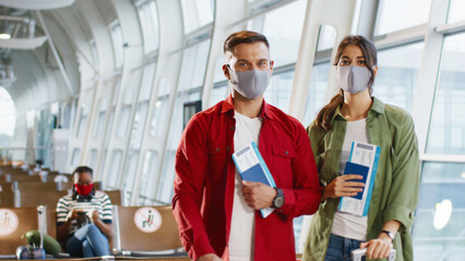 Portrait of the young caucasian cheerful couple wearing protective masks standing at the airport with tickets and passports in hands and smiling to the camera