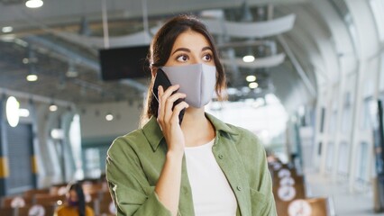 Young pretty smiled caucasian girl with brunette hair wearing protective mask standing in the airport and talking cheerfully on the smartphone