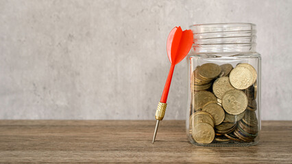Red dart arrow leaning against a glass jar full of gold coins for savings goals and investment success. © MohamadFaizal