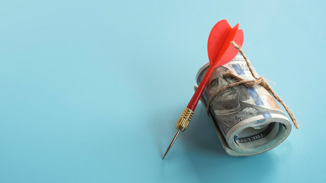 Red dart leaning against a roll of US dollar bills for financial targeting.