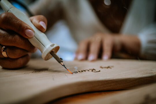 Close-up of a female artisan using a wood burning tool to create intricate designs on a wooden surface in a well-lit workshop setting