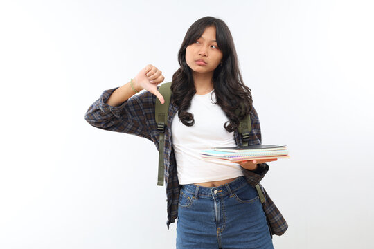 Teenage girl isolated on gray background, schoolgirl with backpack and books, looks unhappy, disappointed, and hates studying, showing thumbs down.