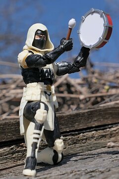 GI JOE Classified series action figure of Storm Shadow, in white hood and kawa-mono mask, beating on small tambourine. Birds nest, tree branches and blie skies in background, spring sunshine.