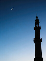 Fototapeta premium Crescent moon over silhouette of minaret against blue sky. Islamic architecture for Ramadan and Eid al-Adha greeting card.