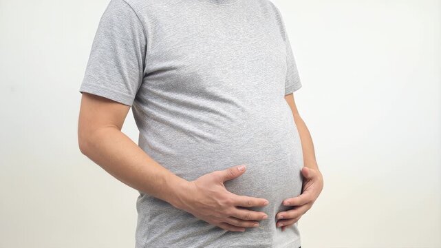 Overweight man in gray t-shirt holding his large belly against white background representing obesity and unhealthy lifestyle choices.