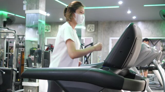 sian woman wearing protective face mask while exercising on treadmill in gym for health safety, air pollution, and PM 2.5 prevention concept