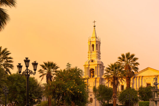 The white volcanic stone facade and ornate bell tower of the Basilica Cathedral of Arequipa glow under a warm sunset sky in Peru.