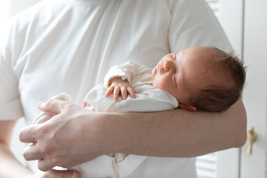 Close-up of a newborn baby sleeping peacefully in the arms of a parent wearing a white shirt. The tender moment, soft natural light, and gentle pose convey warmth, safety, and family bonding.